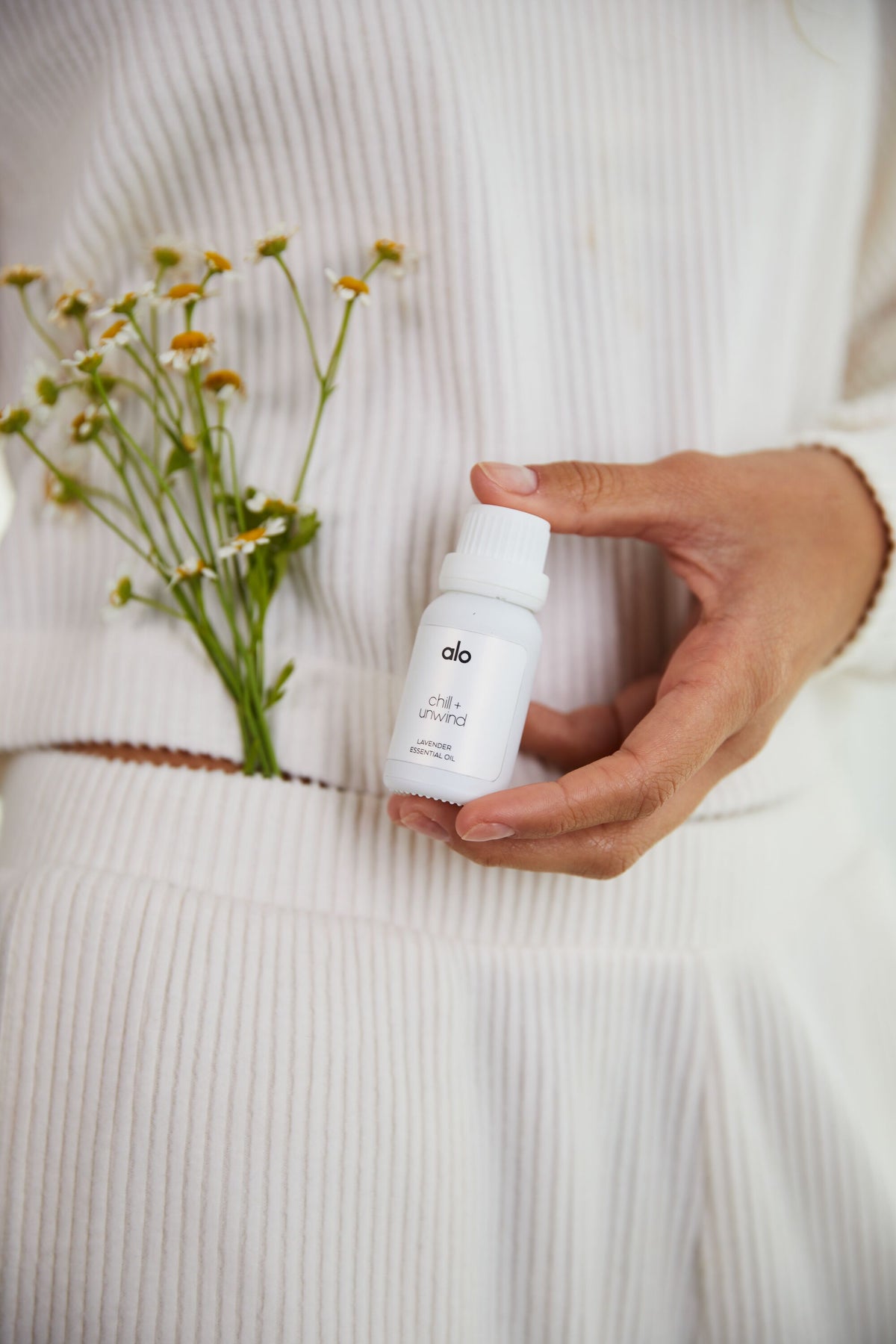 A woman holding a bottle of the Chill + Unwind lavender essential oil while wearing the Muse Shorts and Muse V-Neck Pullover in Ivory with a small bouquet of flowers in her pocket.  