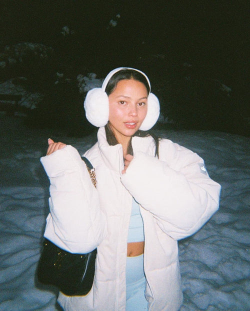 @fiona.bl wearing a light blue workout set with a sherpa puffer jacket on top and faux fur ear muffs while standing on a snow-covered ground at night.